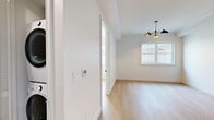 This thoughtfully designed one-bedroom, one-bathroom unit offers a clean, modern aesthetic across its single floor. Light hardwood flooring flows consistently throughout the space, creating a warm and cohesive feel. The open kitchen is a standout feature, showcasing full-height gray shaker-style cabinetry, a neutral tile backsplash, white quartz countertops, stainless steel appliances including a built-in oven and microwave, and a functional island extension that doubles as a dining or prep surface. Recessed lighting illuminates the kitchen workspace effectively. The main living area is bright and airy, benefiting from natural light streaming through windows and accented by a stylish multi-arm pendant light fixture. Crown molding adds a refined architectural touch throughout. The bedroom is a well-proportioned, sun-filled retreat with a large window offering views to the outside, light hardwood floors, and crisp white walls. The bathroom is elegantly appointed with patterned cement-style floor tiles, marble-effect wall panels at half height, a wall-mounted toilet, and a white vanity with matte black hardware, creating a sophisticated and spa-like atmosphere. Overall, the unit combines contemporary design with quality finishes for a polished living environment.