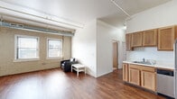 Located at 155 Division Ave N in Grand Rapids, Michigan, this single-floor unit showcases a distinctive urban loft aesthetic rooted in industrial architectural character. Exposed painted brick walls and visible metal ductwork running along the ceiling create a raw, industrial backdrop that is balanced by crisp white walls and warm hardwood flooring throughout the main living areas.

The open living area benefits from multiple windows set within the brick walls, allowing natural light to filter in and offering views of the surrounding cityscape. Soaring ceilings amplify the sense of openness, lending the space a notably airy quality. A defined kitchen features warm wood cabinetry extending to ceiling height, stainless steel appliances including a refrigerator, range, microwave, and dishwasher, along with a sink and ample counter space.

A separate bedroom provides a quieter retreat, finished with carpeted flooring, a window with city views, exposed brick, and a closet. The bathroom includes a wood-toned vanity with a sink, a mirror, and hardwood-style flooring. An entry area with tall ceilings connects the unit's rooms cohesively. Overall, the space combines industrial character with functional, comfortable living in a central Grand Rapids location.