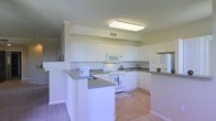 This single-story space centers around a bright, functional kitchen designed for practicality and ease of use. The kitchen features an abundance of white cabinetry that extends to the upper walls, providing generous storage throughout. Granite-style countertops in a neutral tone complement the white cabinetry and run along the perimeter of the kitchen as well as across a prominent peninsula, which serves as both a prep area and a casual dining or social space.

White appliances, including a built-in microwave, electric range, refrigerator, and sink, are seamlessly integrated into the layout, maintaining a cohesive, clean aesthetic. The U-shaped configuration of the countertops and cabinetry maximizes workspace efficiency, while the peninsula adds additional counter space and separates the kitchen from the adjacent living area.

Overhead, a large flush-mount fluorescent light fixture provides bright, even illumination throughout the kitchen. Light-toned flooring adds to the airy feel of the space. Neutral wall tones and crown molding detail along the upper cabinetry contribute to a polished, well-finished interior.