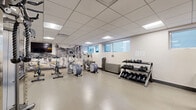 Located at 1745 Avenue Cedar in Montréal, Québec, this fitness facility offers a well-appointed exercise room designed for a complete workout experience. The space features a clean, modern aesthetic with a suspended tile ceiling fitted with recessed panel lighting that ensures bright, even illumination throughout. Durable, easy-to-maintain tile flooring covers the entire floor area, providing a practical surface suited for high-traffic use. A striking black-and-white cityscape mural adorns one wall, adding a dynamic visual element and a nod to Montréal's urban character. Natural light filters in through a row of windows along the upper portion of the walls, complementing the interior lighting. The cardio section is well stocked with treadmills, stationary spin bikes, and elliptical trainers, while a wall-mounted flat-screen television enhances the cardio experience. The strength training area features a multi-function cable machine, a Smith machine with an adjustable bench, and a tiered dumbbell rack with a full range of free weights. Accent walls in bold yellow-orange add energy and visual contrast against the neutral grey and white tones of the room. A mirrored wall section expands the sense of space and supports form-checking during workouts. A small service counter with cabinetry provides additional functionality. Overall, the gym delivers a focused, professional environment for fitness.