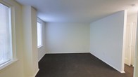 This single-level space presents a straightforward, minimalist design centered around a well-proportioned room finished with clean white walls and a smooth white ceiling that together create a bright, open atmosphere. Dark carpeting covers the floor throughout, offering a grounding contrast to the lighter tones of the walls and ceiling. A window positioned along one wall allows natural light to filter into the space, contributing to an airy feel despite the room's simple layout. White baseboards frame the perimeter of the room, adding a subtle but clean finishing detail. The overall condition of the space appears well-maintained, with neutral finishes that provide a versatile canvas suitable for a variety of uses or furnishing styles. The uncluttered design and neutral color palette make the room highly adaptable, whether intended for residential or professional use.
