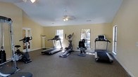 Located at 1398 S. University Drive in Ladson, South Carolina, this single-floor fitness center offers a dedicated space for cardiovascular exercise and general wellness. The room is finished with warm yellow walls and features durable, carpet-tiled flooring well-suited for a high-traffic workout environment. Multiple windows allow natural light to fill the space, creating a bright and welcoming atmosphere that enhances the overall workout experience. A ceiling fan with an integrated light fixture is centrally mounted, providing consistent air circulation throughout the room. The facility is equipped with a variety of cardio machines, including multiple treadmills and an elliptical trainer, as well as a stationary bike, accommodating a range of fitness preferences and routines. The layout is thoughtfully arranged to allow comfortable spacing between equipment, ensuring ease of movement and safe use. Recessed vents support climate control, keeping the environment comfortable year-round. Clean white baseboards and a well-maintained interior reflect an organized, functional facility designed to support an active lifestyle.