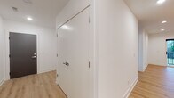 This single-floor home showcases a clean, modern aesthetic defined by crisp white walls, light hardwood-style flooring, and an open, airy layout that flows seamlessly from room to room. The kitchen is a standout feature, offering white shaker cabinetry, light quartz countertops, a full suite of stainless steel appliances — including a refrigerator, range, microwave, and dishwasher — and a large central island with a built-in sink. Edison-style pendant lights hang above the island, adding a warm, contemporary touch to the space. Recessed lighting and natural light from nearby windows keep the area bright and inviting. The living areas continue the open feel, with ceiling fans providing comfort and windows that frame outdoor views. A dedicated walk-in closet offers generous built-in shelving and hanging space, providing excellent storage. The bathroom features a white vanity with a single sink, a large mirror with overhead lighting, and hardwood-style flooring that ties cohesively with the rest of the home. Hallways are wide and well-lit, featuring tall, flush-mount doors with polished chrome hardware that add a refined, modern detail. Overall, the space presents a cohesive, contemporary design with thoughtful finishes throughout.