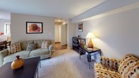 This single-floor residence offers a thoughtfully organized layout that balances comfort and functionality throughout. The living room serves as the heart of the home, featuring soft wall-to-wall carpeting, neutral gray-toned walls, and warm ambient lighting that creates an inviting atmosphere. Framed artwork and a curated mix of seating arrangements add character to the space, which flows openly toward the rest of the unit.

The galley-style kitchen is efficiently designed with white cabinetry, a double-basin sink, laminate countertops, and wood-look flooring. Black appliances, including a refrigerator and electric range, provide a sleek contrast to the bright cabinetry. The kitchen connects to a hallway that leads to additional rooms, including a walk-in closet area.

A dedicated dining nook is brightened by a window with sheer white curtains and wood blinds, filling the area with natural light.

The unit includes two bedrooms, both carpeted and fitted with ample closet space. The primary bedroom features multiple windows with dark curtain panels, a textured ceiling, and generous room for furnishings. The second bedroom is quieter in scale but equally functional, with a built-in closet and cozy feel.

The full bathroom offers a white vanity, combination tub and shower, and wood-look tile flooring. A dedicated utility closet houses the water heater and wire shelving for additional storage.