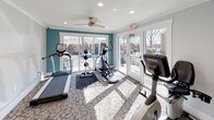 This fitness center is a bright, thoughtfully designed workout facility that provides residents with a comprehensive range of exercise options. The space is characterized by large sliding glass windows and doors that line the walls, flooding the interior with natural light and offering open views of the surrounding community's landscaping and buildings. Teal accent walls add a vibrant, energizing touch to the otherwise neutral gray and white color palette, while patterned carpeting covers the floor throughout.

The cardio area is well-appointed with True-brand upright and recumbent stationary bikes, a treadmill, and an elliptical trainer, all positioned to take advantage of the outdoor views. A dedicated strength training section features a multi-station cable machine along with a weight bench and additional resistance equipment, providing a full-body workout experience. Medicine balls and supplementary accessories are also available.

Overhead ceiling fans help maintain comfortable airflow, and recessed lighting ensures the space remains well-lit. A wall-mounted mini-split air conditioning unit provides climate control. Amenities include a dual water fountain station with a bottle-filling feature and a wall-mounted television. Posted fitness center rules are displayed for user reference. The abundance of windows and glass doors creates an open, airy ambiance that makes workouts feel connected to the outdoors.