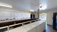Located at 221 Smilax in Vista, this single-floor community laundry facility is designed to provide a practical and efficient laundry experience. The space is outfitted with a generous row of commercial-grade top-loading washing machines aligned along a central counter, complemented by a full bank of stacked dryers lining the back wall, offering substantial capacity for residents. The flooring throughout is a rich, dark wood-tone laminate that adds a clean, modern touch to the utilitarian space. Overhead fluorescent lighting ensures the room is well-lit and functional at any hour. A folding counter runs along one side, providing a convenient surface for sorting and folding laundry. The facility also includes both a snack vending machine and a beverage vending machine, adding everyday convenience for users. Informational postings and laundry guidelines are displayed on the walls for easy reference. A large glass sliding door at the entrance floods the space with natural light and opens directly to a landscaped outdoor area with mature trees and a paved walkway, creating a pleasant, airy atmosphere. A blue entrance mat welcomes visitors at the threshold. The overall layout is straightforward and well-organized, making this laundry facility a highly functional community amenity.