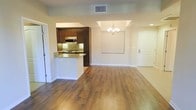 This single-floor space showcases an open-concept design that seamlessly connects the kitchen, dining, and living areas. Warm hardwood-style flooring runs throughout, lending a cohesive and inviting feel to the entire space. The kitchen is defined by rich dark wood cabinetry, granite countertops with subtle veining, and a mosaic tile backsplash that adds visual texture. A peninsula-style counter extends into the open area, providing additional prep space and a natural dividing point between the kitchen and the rest of the living space. Stainless steel appliances, including a built-in microwave, complement the kitchen's modern aesthetic. The dining area is highlighted by an elegant chandelier suspended from a tray ceiling detail, adding a refined architectural touch. Adjacent to the dining space, a recessed niche in the wall offers a designated area suited for a large display or mounted screen. Natural light filters into the space, while neutral-toned walls provide a clean, versatile backdrop. A paneled entry door with a sidelite is visible, offering a welcoming transition point into the unit. Overall, the space presents a functional and polished interior with thoughtful design details throughout.