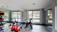 This single-floor fitness studio is designed to support a well-rounded workout experience in a clean, modern environment. The space features multiple stationary cycling bikes positioned to take full advantage of the large windows that line the walls, offering views of the surrounding greenery and filling the room with abundant natural light. Roller shades on each window allow for adjustable light control during workouts. A collection of exercise balls in varying sizes adds versatility for strength and balance training. The floor is finished with durable hardwood-style planking, providing both a practical and polished aesthetic underfoot. Full-length mirrors are mounted along one wall, expanding the sense of space and allowing users to monitor their form during exercise. The walls are finished in a neutral grey tone, contributing to a focused and energizing atmosphere. Recessed lighting supplements the natural light, ensuring the space remains well-lit at all hours. The overall design is functional and purposeful, with an open layout that accommodates group fitness sessions or individual training. The combination of natural light, mirrored walls, and modern finishes gives this fitness studio a bright, motivating ambiance.