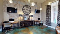 This distinctive single-floor space makes a bold design statement through its thoughtful fusion of industrial and eclectic styles. The centerpiece of the main room is a dramatic exposed brick wall, which provides a warm, textured backdrop for an oversized wrought-iron Roman numeral clock that commands attention. Two flat-screen televisions are mounted at either side of the wall, complementing a dark wood media console on industrial caster wheels.

The flooring is a true standout feature — acid-stained concrete in rich swirling tones of teal, gold, and deep brown creates a one-of-a-kind, almost artistic surface that anchors the entire aesthetic of the space. Pendant globe lights hang from above, casting a warm ambient glow that enhances the room's inviting character.

A functional workspace area is thoughtfully integrated, featuring a wooden desk with a rolling office chair, ideal for productive use within the space. Decorative table lamps with dark shades add warmth and symmetry to the console arrangement, while a framed mirror and curated accessories introduce refined touches. Patterned drapery panels frame a window, softening the room's harder industrial elements. Overall, the space exudes a confident, creative personality with a cohesive blend of rugged textures and refined details.