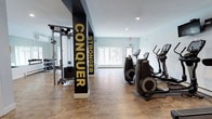 This fitness center offers a comprehensive workout environment designed to support a wide range of exercise routines. The space is spread across a single floor and features a light, airy atmosphere thanks to light blue and white walls, recessed lighting, and windows that invite natural light into the interior.

Structural columns serve a dual design purpose, wrapped in bold navy and black panels displaying motivational words such as "Conquer," "Stronger," "Challenge," "Energize," and "Workout" in gold lettering. These elements give the space a dynamic, gym-forward identity while clearly defining different zones throughout the floor.

The cardio section is well-appointed with multiple elliptical machines, treadmills, and upright and recumbent stationary bikes positioned to allow ample space between equipment. A dedicated strength training area features a Life Fitness cable machine, a weight bench, and a rack of dumbbells for free weight exercises. Full-length mirrors line portions of the wall, enhancing spatial depth and supporting proper form during workouts.

Durable wood-look flooring extends throughout, offering both a polished aesthetic and a practical surface for exercise. The overall layout is open and uncluttered, allowing users to move freely between equipment zones.