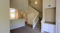 This two-story residence showcases a thoughtfully designed open-concept layout that maximizes the sense of space throughout. The main level features carpeted flooring that extends through the living area and up the staircase, creating a cohesive and comfortable feel underfoot. Vaulted ceilings rise dramatically above the main living space, adding an airy, expansive quality to the interior.

A carpeted staircase with white-painted wood railings and a dark wood handrail connects the two floors, serving as a central architectural element. Beneath the staircase, a classic six-panel closet door provides practical storage. Adjacent to the living area, a raised countertop breakfast bar delineates the kitchen space, which is visible in the background alongside a window that draws in natural light.

Built-in niche shelving near the entryway offers display or storage opportunities. A hallway beyond the living space leads to additional rooms, suggesting a well-distributed floor plan. The neutral warm-toned walls throughout create a welcoming ambiance, while recessed vents and ceiling-mounted light fixtures contribute to the home's functional design.