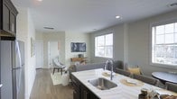 This single-floor space showcases a thoughtfully designed open-concept layout that integrates the kitchen and living area into one cohesive, functional environment. The kitchen is anchored by a substantial island topped with a light-toned striped countertop, featuring a deep stainless steel double-basin sink with a sleek pull-down faucet. Dark cabinetry beneath the island provides a striking contrast to the lighter surfaces above. Recessed lighting in the smooth ceiling complements the natural light that filters through a large window dressed with white horizontal blinds in the living area. The living space is furnished with contemporary gray seating, a flat-screen television mounted above a console table, and a soft area rug over hardwood-style flooring that runs throughout. Neutral, light gray walls create a calm and cohesive ambiance across the entire space. A doorway visible along one wall suggests access to additional rooms, contributing to the overall sense of a well-connected and efficiently arranged floor plan.