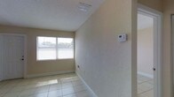 This single-story space presents a straightforward and functional interior designed for versatility. The room showcased in the images features large format, light-toned tile flooring that reflects natural light, lending a bright and open feel to the space. The walls carry a subtle textured finish in a warm neutral tone, complemented by clean white baseboards that add a crisp, polished contrast throughout.

A window fitted with horizontal blinds allows natural light to filter in while maintaining privacy, contributing to the room's airy atmosphere. Overhead, a textured ceiling houses a centrally positioned ventilation grille, supporting effective airflow throughout the space. A wall-mounted programmable thermostat provides convenient and precise climate control, adding to the room's practical functionality.

The overall ambiance is clean, neutral, and well-maintained, making the space adaptable to a range of uses. The combination of durable tile flooring, climate management features, and natural light creates an environment that is both comfortable and easy to maintain — suitable for residential or light professional use.
