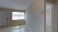 This single-story space presents a straightforward and functional interior designed for versatility. The room showcased in the images features large format, light-toned tile flooring that reflects natural light, lending a bright and open feel to the space. The walls carry a subtle textured finish in a warm neutral tone, complemented by clean white baseboards that add a crisp, polished contrast throughout.

A window fitted with horizontal blinds allows natural light to filter in while maintaining privacy, contributing to the room's airy atmosphere. Overhead, a textured ceiling houses a centrally positioned ventilation grille, supporting effective airflow throughout the space. A wall-mounted programmable thermostat provides convenient and precise climate control, adding to the room's practical functionality.

The overall ambiance is clean, neutral, and well-maintained, making the space adaptable to a range of uses. The combination of durable tile flooring, climate management features, and natural light creates an environment that is both comfortable and easy to maintain — suitable for residential or light professional use.