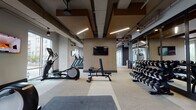 Located at 500 W Trade Street in the heart of Charlotte, NC, this contemporary fitness center is a well-appointed, single-floor facility designed to support a full range of workout routines. The space is divided into distinct training zones, each thoughtfully equipped and finished with a cohesive modern aesthetic. Warm wood-toned accent panels and rich ceiling coffers contrast against exposed structural elements and sleek linear lighting, creating an industrial-modern atmosphere throughout.

The cardio zone features multiple treadmills, elliptical trainers, and additional aerobic equipment, all positioned alongside expansive floor-to-ceiling windows that invite abundant natural light and offer open views of the surrounding Charlotte cityscape. Wall-mounted flat-screen televisions are positioned throughout for entertainment during workouts.

The strength training area includes a full rack of dumbbells, a Smith machine, a hyperextension bench, and various resistance machines from Precor and other commercial-grade brands. Rubber flooring and padded mat areas provide safe, dedicated space for free weight exercises.

A separate functional training room, enclosed by glass partition walls, provides a versatile open floor plan ideal for group fitness, stretching, or boxing, with a heavy bag visible inside. Floor-to-ceiling mirrors amplify the sense of space and support form-conscious training. The facility is finished with neutral-toned carpeting and ceiling fans throughout, ensuring comfort across all zones.