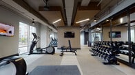 Located at 500 W Trade Street in the heart of Charlotte, NC, this contemporary fitness center is a well-appointed, single-floor facility designed to support a full range of workout routines. The space is divided into distinct training zones, each thoughtfully equipped and finished with a cohesive modern aesthetic. Warm wood-toned accent panels and rich ceiling coffers contrast against exposed structural elements and sleek linear lighting, creating an industrial-modern atmosphere throughout.

The cardio zone features multiple treadmills, elliptical trainers, and additional aerobic equipment, all positioned alongside expansive floor-to-ceiling windows that invite abundant natural light and offer open views of the surrounding Charlotte cityscape. Wall-mounted flat-screen televisions are positioned throughout for entertainment during workouts.

The strength training area includes a full rack of dumbbells, a Smith machine, a hyperextension bench, and various resistance machines from Precor and other commercial-grade brands. Rubber flooring and padded mat areas provide safe, dedicated space for free weight exercises.

A separate functional training room, enclosed by glass partition walls, provides a versatile open floor plan ideal for group fitness, stretching, or boxing, with a heavy bag visible inside. Floor-to-ceiling mirrors amplify the sense of space and support form-conscious training. The facility is finished with neutral-toned carpeting and ceiling fans throughout, ensuring comfort across all zones.