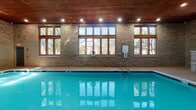 Located at 2705 Pontiac Dr in Aurora, Illinois, this indoor pool facility offers a well-appointed aquatic space designed with both function and aesthetics in mind. The centerpiece is a generously sized rectangular swimming pool with vivid turquoise water, trimmed with blue tile accents along the pool's edge and clearly marked depth indicators along the deck.

The architecture blends natural materials throughout, with textured stone block walls rising to meet a warm, rich wood-paneled ceiling fitted with recessed lighting that illuminates the space evenly. Large wood-framed windows line the walls on multiple sides, flooding the interior with natural light and providing views of the surrounding mature trees and neighborhood exterior.

The pool deck is finished in light-toned concrete, offering ample circulation space around the perimeter. A hot tub or spa feature is visible in one corner, also clad in matching stone. Entry into the pool is facilitated by stainless steel handrails, and curved stone steps near the entrance area add a decorative architectural detail. Safety signage, including a no-diving notice, is posted prominently. The facility includes a secured entry door and appears well-maintained, making it a practical and visually appealing amenity for its community in Aurora.