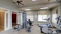 Located at 101 Crossing Dr in Millbrook, Alabama, this fitness center offers a well-equipped, single-floor space designed to support a variety of workout routines. The room is finished with neutral gray walls and carpeted flooring, creating a clean and focused environment for exercise. Multiple windows dressed with valances and horizontal blinds allow generous natural light to fill the space, while a ceiling fan overhead promotes air circulation alongside recessed fluorescent lighting panels.

The fitness area is outfitted with a multi-station cable weight machine, providing users with a versatile strength-training option capable of targeting multiple muscle groups. Alongside the strength equipment, cardio machines — including a treadmill and an elliptical trainer — round out the facility's offerings. A framed informational notice is mounted on the wall, likely detailing fitness center rules or guidelines for users.

A doorway visible in the background leads to an adjacent area, suggesting additional amenity spaces nearby. The overall ambiance of the room is practical and inviting, with ample space between equipment for comfortable use. The combination of natural light, ceiling ventilation, and well-maintained equipment makes this fitness center a functional and welcoming space for residents or guests seeking an accessible on-site workout facility.