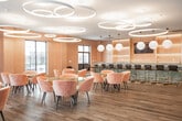 Located at 695 Merrick Ave in Westbury, New York, this thoughtfully designed single-floor amenity lounge offers a sophisticated and welcoming environment with a cohesive contemporary aesthetic. The space is unified by warm wood-plank flooring that flows seamlessly across multiple functional zones, complemented by soft peach and warm neutral wall tones that create an inviting atmosphere throughout.

The dining area is a standout feature, anchored by sculptural round tables paired with plush velvet chairs in warm terracotta and blush tones. Overhead, a dramatic cluster of interlocking circular ring pendant lights serves as a striking focal point, casting a warm, layered glow across the space. Large black-framed windows line the walls, drawing in abundant natural light and offering views of the surrounding exterior.

A lounge zone provides comfortable seating for relaxation or casual gatherings, featuring a teal velvet sofa set atop a bold geometric patterned area rug in shades of blue and white. This area is further defined by a paneled accent wall with integrated lighting and direct access to the outdoors via French doors.

A bar counter finished in vibrant green marble with gold-framed barstools adds a glamorous touch to the space. An additional media lounge area includes a wall-mounted television, ample seating, and patterned accent wallpaper, rounding out this versatile and elegantly appointed communal space.