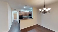 This single-floor residence offers a well-designed, open-concept layout that seamlessly connects the kitchen and dining areas, creating a cohesive and functional living environment. The rich, dark hardwood flooring extends throughout the main living spaces, providing a warm and grounded aesthetic that ties the rooms together.

The kitchen is defined by a raised breakfast bar peninsula with dark granite countertops, offering both additional prep space and a natural partition from the dining area. Ample rich, medium-toned wood cabinetry lines the walls, providing generous storage, while black appliances — including a refrigerator, range, and built-in microwave — lend a sleek, uniform look. Recessed lighting overhead ensures the kitchen is well-lit and practical.

The adjacent dining area is accentuated by an elegant wrought-iron chandelier, adding a touch of traditional charm to the space. A matching wall sconce nearby reinforces the cohesive design language. Arched doorways lead into a hallway, hinting at the additional rooms beyond and contributing an architectural detail that softens the overall aesthetic. The neutral, warm-toned walls throughout complement the darker flooring and cabinetry, resulting in a balanced and inviting atmosphere.