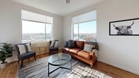 River's Edge is a two-bedroom, two-bathroom residence that combines contemporary design with sweeping views of the river and surrounding cityscape. The open-concept living area is anchored by large windows on multiple walls, flooding the space with natural light and framing impressive outdoor vistas. Hardwood-style flooring runs throughout the main living area, complementing a bright, neutral color palette.

The kitchen is a standout feature, outfitted with white cabinetry, gray countertops, stainless steel appliances, and a spacious island with bar seating for four. Pendant lighting adds a polished finish, while an adjacent dining area benefits from continued river views. The living room extends the open layout, offering generous space and additional corner windows that capture broad panoramic scenery.

Both bedrooms feature plush carpeting, ceiling fans, and windows with outdoor views. The primary bedroom connects to a private bathroom with a glass-enclosed walk-in shower and gray stone countertops. The second bedroom includes mirrored sliding closet doors and flexible space suitable as a guest room or home office. The second full bathroom features a soaking tub and a matching stone-topped vanity, maintaining the home's cohesive, clean aesthetic throughout.