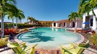 Located at 15270 Ballast Point Dr in Fort Myers, Florida, this single-story community amenity area offers a resort-style outdoor experience designed for relaxation and social gatherings. At its centerpiece is a generously sized freeform swimming pool with clear turquoise water, surrounded by a broad brick-paver deck lined with lounge chairs for sunbathing under the abundant Florida sunshine. Towering palm trees and lush tropical plantings frame the pool area, creating a vibrant, resort-like ambiance throughout.

Adjacent to the pool, a covered outdoor pavilion provides a comfortable shaded retreat. Ceiling fans maintain airflow throughout the space, and a wall-mounted television makes it ideal for entertaining. The pavilion features multiple wicker dining tables and chairs with colorful striped cushions, along with a lounge seating area. A bar counter with bar seating and access to a kitchenette area with a refrigerator adds further convenience for gatherings.

Open-air columns and wide archways connect the pavilion to the surrounding pool deck, offering unobstructed views of the pool and tropical landscape. The brick-paver flooring flows seamlessly from the covered areas to the outdoor spaces, unifying the entire amenity area into a cohesive, inviting environment well-suited to the Southwest Florida lifestyle.