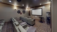 This dedicated screening theater delivers a refined cinematic experience within a thoughtfully designed, intimate setting. The room is arranged with multiple rows of plush, light gray lounge chairs, each equipped with an attached dark wood side tray — ideal for snacks and beverages during a screening. The seating is oriented toward a large, wall-mounted projection screen framed in black, providing a commanding focal point at the front of the room. A ceiling-mounted projector ensures crisp, high-quality image projection, while in-wall speakers positioned throughout the space deliver immersive surround sound.

The interior design draws on classic cinema nostalgia, with decorative oversized film reels mounted on the walls serving as striking, thematic accents. Warm sconce lighting along the walls creates a cozy, dimly lit ambiance suited for movie viewing, complemented by recessed ceiling lights for general illumination. Rich, patterned carpeting covers the floor, adding warmth and acoustic absorption to the space.

Near the entrance, a traditional popcorn machine on a wheeled cart adds a charming, authentic movie-going touch, alongside a small round café table. Wood-trimmed crown molding and dark baseboards lend a polished finish to the overall design, making this theater a well-appointed venue for private screenings and group entertainment.
