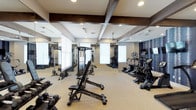 This fitness center offers a comprehensive workout environment designed to accommodate a wide range of exercise routines. The space is defined by its distinctive architectural ceiling detail, featuring dark-stained exposed wood beams that add a refined, rustic character to the otherwise modern interior. Recessed lighting and ceiling fans ensure the space remains well-lit and comfortable during use.

Floor-to-ceiling mirrors line one wall, visually expanding the space and allowing users to monitor their form during workouts. Large windows with blinds bring in natural light while maintaining privacy. The flooring is a durable, light-toned carpet suited for gym use.

On the cardio side, a row of treadmills and elliptical machines is aligned along the mirrored wall, each equipped with individual digital consoles. The strength training area features a multi-function cable machine, an adjustable weight bench, and a full rack of dumbbells, providing options for both isolation and compound movements. The equipment selection supports users of varying fitness levels, from beginners to more advanced athletes.

Overall, the fitness center strikes a balance between functionality and aesthetic appeal, creating an inviting environment that encourages regular use.