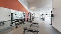 This expansive fitness center offers a comprehensively outfitted exercise facility spread across a single level, thoughtfully divided into distinct workout zones. The space is characterized by clean, modern design with crisp white walls, recessed lighting, and an open layout that promotes ease of movement throughout.

The cycling studio occupies one section of the facility, featuring hardwood-style flooring and multiple stationary bikes arranged for group fitness sessions. A wall-mounted flat-screen television provides entertainment, while several windows along the perimeter bring in natural light and outdoor views.

An adjoining cardio and strength zone features durable rubber flooring and houses a variety of machines, including treadmills, ellipticals, cable machines, and multi-station strength equipment. Mounted televisions provide visual engagement during workouts.

A dedicated free weights area is distinguished by a bold terracotta accent wall and full-length mirrors that visually expand the space and aid in form monitoring. This zone includes a comprehensive dumbbell rack, weight benches, medicine balls, and a power rack, catering to a full range of strength training needs.

Throughout the facility, soaring ceilings enhance the sense of openness, while the well-planned zoning ensures that multiple workout styles can be pursued simultaneously without congestion.
