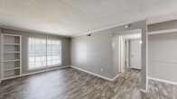This single-floor unit presents a cohesive, neutral aesthetic defined by gray-toned walls, crisp white trim, and wood-look plank flooring that flows consistently throughout the space. The layout, as revealed by the floor plan model, is well-organized and makes efficient use of its footprint, connecting a main living area, secondary room, kitchen, and bathroom in a logical arrangement.

The main living area is generously proportioned, featuring a textured popcorn ceiling, crown molding, and a large window fitted with horizontal blinds that allows ample natural light. A built-in shelving unit flanks the window, adding both character and practical storage. An adjacent open area connects seamlessly to the front entry, creating an easy flow between spaces.

The kitchen runs along a galley-style corridor and is equipped with dark cabinetry, light-toned countertops, and black appliances including a range and refrigerator. Overhead cabinetry maximizes storage, and under-cabinet lighting provides functional task illumination.

The bedrooms continue the consistent flooring and gray palette. One bedroom features a built-in closet with multi-tiered shelving, while windows in each room invite natural light. The bathroom includes a full tub and shower combination with large-format gray tile surround, complementing the unit's overall color scheme.