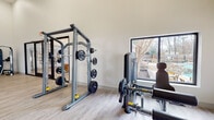 Located at 5620 Wood Run Blvd in Henrico, Virginia, this fitness center is a well-appointed, single-floor facility designed to support a wide range of workout routines. The space features soaring ceilings that lend an open, airy atmosphere, complemented by neutral white walls and light-toned luxury vinyl plank flooring throughout.

The cardio zone is equipped with TRUE-brand treadmills, a stair climber, and a stationary bike, positioned near large picture windows that frame views of the outdoor courtyard and surrounding landscaping, allowing natural light to fill the space during workouts. A wall-mounted television provides entertainment for cardio sessions.

The strength training area includes a full power rack loaded with weight plates, a multi-function cable machine, and a dedicated functional training rig complete with suspension straps, resistance bands, and battle ropes. A well-stocked free weights rack holds an organized assortment of dumbbells, kettlebells, and medicine balls for varied training needs.

The facility opens via sleek, floor-to-ceiling black-framed glass sliding doors, connecting seamlessly to an adjacent lounge area and providing a modern, polished aesthetic. A wall-mounted TV and a small accent table round out the space, making it both functional and inviting for residents seeking a comprehensive on-site fitness experience.