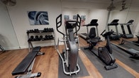 This fitness center is a comprehensively outfitted exercise facility spread across a single level, designed to accommodate a wide range of training needs. The main workout area features warm-toned hardwood-style flooring, recessed lighting, and ceiling-mounted fans that contribute to a comfortable and functional environment. A full-length wall mirror visually expands the space while allowing users to monitor their form during exercise.

The cardio zone is well-appointed, featuring a treadmill, elliptical trainer, recumbent bike, and a Concept2 rowing machine — providing options for both low-impact and high-intensity cardiovascular workouts. A multi-station functional cable trainer anchors the strength training area, complemented by a tiered dumbbell rack stocked with a range of free weights and a flat workout bench.

A separate adjoining room provides dedicated space for flexibility and functional training, equipped with stability balls in multiple sizes and storage shelving for accessories. A paper towel dispenser and waste bin are conveniently placed near the entrance, supporting cleanliness and hygiene. Motivational sports artwork adorns the walls throughout, adding an energetic aesthetic to the space. Staircase access is visible, indicating the facility's integration within a larger building.