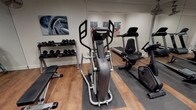 This fitness center is a comprehensively outfitted exercise facility spread across a single level, designed to accommodate a wide range of training needs. The main workout area features warm-toned hardwood-style flooring, recessed lighting, and ceiling-mounted fans that contribute to a comfortable and functional environment. A full-length wall mirror visually expands the space while allowing users to monitor their form during exercise.

The cardio zone is well-appointed, featuring a treadmill, elliptical trainer, recumbent bike, and a Concept2 rowing machine — providing options for both low-impact and high-intensity cardiovascular workouts. A multi-station functional cable trainer anchors the strength training area, complemented by a tiered dumbbell rack stocked with a range of free weights and a flat workout bench.

A separate adjoining room provides dedicated space for flexibility and functional training, equipped with stability balls in multiple sizes and storage shelving for accessories. A paper towel dispenser and waste bin are conveniently placed near the entrance, supporting cleanliness and hygiene. Motivational sports artwork adorns the walls throughout, adding an energetic aesthetic to the space. Staircase access is visible, indicating the facility's integration within a larger building.