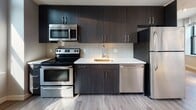 Located at 23 Central in Lynn, Massachusetts, this modern single-floor unit offers a thoughtfully designed layout with contemporary finishes throughout. The open-concept living and kitchen area serves as the heart of the space, featuring rich hardwood-style flooring that flows seamlessly from room to room. The kitchen is outfitted with sleek dark cabinetry, light countertops, a white subway tile backsplash, and a full suite of stainless steel appliances including a refrigerator, electric range, built-in microwave, and dishwasher — providing both style and functionality. Track lighting above accents the space effectively. The living area is generously proportioned, benefiting from abundant natural light streaming through multiple black-framed windows fitted with horizontal blinds. These windows frame views of Lynn's surrounding urban architecture, including the classic brick buildings of the downtown area. The bedrooms continue the clean, neutral aesthetic with soft gray walls, hardwood-style floors, and well-placed windows that bring in natural light and city views. The bathroom is finished with large-format tile flooring, a white vanity, and a full bathtub and shower surround. Overall, the unit presents a clean, modern ambiance well-suited to urban living in a vibrant city setting.