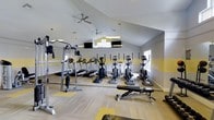 This fitness center offers a thoughtfully designed workout environment within a single-floor layout. The space is defined by striking vaulted ceilings that create an open, airy atmosphere, complemented by multiple ceiling fans that ensure comfortable airflow during workouts. Natural light streams in through several windows, including a set of upper-level windows positioned near the peak of the vaulted ceiling, brightening the space throughout the day.

The interior features light hardwood-style flooring accented by yellow floor mats strategically placed beneath equipment zones. The walls carry a two-tone color scheme with a yellow horizontal stripe against white, adding a energetic and motivating visual element to the environment.

The cardio area is well-appointed with a row of treadmills, elliptical trainers, and a stationary cycle, all aligned along a mirrored wall that visually expands the space and allows users to monitor their form. The strength training section includes a multi-functional cable and weight machine alongside an adjustable workout bench, providing options for a full-body training session.

Wall-mounted televisions are also present, offering entertainment during workouts. Overall, the fitness center combines practical equipment, bright aesthetics, and smart spatial design to deliver a functional and inviting exercise facility.