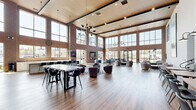 Located at 110 Woodcrest Dr in Cherry Hill, NJ, this expansive single-floor living room is a striking communal space defined by its dramatic double-height ceilings and industrial-modern aesthetic. Exposed brick walls contrast beautifully with sleek floor-to-ceiling steel-framed windows that flood the interior with abundant natural light and offer open views of the surrounding community.

Rich hardwood flooring flows seamlessly throughout, anchoring multiple distinct zones within the open-plan layout. A central lounge area features clusters of contemporary swivel armchairs arranged around low tables, ideal for casual gatherings. Adjacent to this, a bar-height communal table with modern seating provides a more structured setting for socializing or working. A dedicated seating nook showcases a vivid large-scale koi fish painting, adding a bold artistic focal point to the space.

Along one wall, built-in booth seating with leather cushions and gooseneck wall sconces creates a cozy, café-style dining alcove. The space also includes a fireplace with a mounted flat-screen television, further enhancing its comfort and versatility. A welcoming entry area features a glass-railed staircase, a vibrant teal accent wall, and curated décor. A warm wood-paneled ceiling with recessed lighting and statement pendant chandeliers completes the refined yet inviting atmosphere.