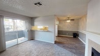 Located at 7000 S Paradise Rd in Las Vegas, Nevada, this single-floor residence offers a practical and comfortable layout designed for everyday living. The main living area features plush carpeting and neutral gray walls, creating a calm and welcoming atmosphere. A sliding glass door floods the space with natural light and provides direct access to a private covered patio, perfect for enjoying the Nevada climate. A pass-through counter with a sink connects the living area to the kitchen, enhancing the open feel of the floor plan. The adjoining room, visible through the pass-through, features wood-look flooring and a ceiling fan, offering versatile space for dining or relaxing. The kitchen is well-appointed with white cabinetry, ample counter space, and a full suite of white appliances, including a refrigerator, range, and dishwasher. A tray ceiling with warm lighting adds a subtle architectural detail overhead, while a window above the sink brings in natural light. Wood-look flooring runs throughout the kitchen and connecting areas, tying the space together with a clean, cohesive look. The textured ceilings and crisp white trim throughout lend a classic, tidy finish to the overall interior.