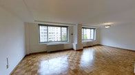 This single-floor space showcases a timeless interior design defined by warm, patterned parquet hardwood flooring that adds classic character throughout the open living area. The room is bathed in abundant natural light, courtesy of multiple large windows that frame views of the surrounding urban cityscape, including neighboring mid-rise buildings and mature trees. The windows are fitted with baseboard heating units, ensuring year-round comfort. The crisp white walls and smooth white ceiling create a clean, airy aesthetic that complements the warmth of the wood floors. The open layout provides a generous sense of space, offering flexibility for a variety of furniture arrangements. The architectural detailing is straightforward and refined, with subtle wood baseboards framing the perimeter of the room. The overall ambiance is bright, neutral, and welcoming, making it a versatile canvas well-suited for both residential and professional use. The combination of natural light, classic flooring, and urban views gives this space a distinct and enduring appeal.