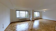 This single-floor space showcases a timeless interior design defined by warm, patterned parquet hardwood flooring that adds classic character throughout the open living area. The room is bathed in abundant natural light, courtesy of multiple large windows that frame views of the surrounding urban cityscape, including neighboring mid-rise buildings and mature trees. The windows are fitted with baseboard heating units, ensuring year-round comfort. The crisp white walls and smooth white ceiling create a clean, airy aesthetic that complements the warmth of the wood floors. The open layout provides a generous sense of space, offering flexibility for a variety of furniture arrangements. The architectural detailing is straightforward and refined, with subtle wood baseboards framing the perimeter of the room. The overall ambiance is bright, neutral, and welcoming, making it a versatile canvas well-suited for both residential and professional use. The combination of natural light, classic flooring, and urban views gives this space a distinct and enduring appeal.