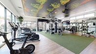 Located in Phoenix, AZ, this expansive fitness center offers a generously sized, single-floor layout designed to accommodate a wide variety of workout routines. One of the most distinctive architectural features is the dramatic ceiling mural depicting a lush tree canopy, adding an immersive, nature-inspired ambiance to the space. Large, floor-to-ceiling glass garage doors line one wall, flooding the interior with natural light and providing open views of the surrounding community.

The flooring transitions between smooth wood-look planks and green turf zones, clearly delineating functional training areas. The turf section serves as the primary functional fitness area, housing a multi-station cable machine, a heavy punching bag, a large tractor tire for functional training, and a trampoline-style training disc. Ceiling-mounted fans ensure proper air circulation throughout.

The cardio zone features a row of treadmills with individual screens positioned alongside the glass walls, complemented by ellipticals and stationary bikes. The strength training area includes a robust selection of selectorized weight machines, free weights with a full rack, and benches, all enhanced by mirrored walls that amplify the sense of space. Wall-mounted flat-screen TVs are positioned throughout for entertainment. The overall design is modern, energetic, and purpose-built for comprehensive fitness use.