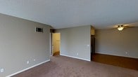 This single-floor space presents a straightforward and functional layout with a neutral, understated aesthetic throughout. The main living area is finished with wall-to-wall carpet in a warm beige tone, providing a comfortable underfoot feel, while the walls are painted in a soft gray that lends a clean, modern look to the interior.

The textured popcorn ceiling adds a classic residential character to the space. A wall-mounted HVAC unit is positioned near the upper left corner of the main room, offering in-unit climate control. White baseboards provide a clean contrast against the gray walls and carpet.

Visible through an open doorway, a walk-in closet offers generous storage space with carpeted flooring and a hanging rod, making it a practical feature for organizing belongings. To the right, a transition to hard flooring marks the beginning of a kitchen area, where upper cabinetry in a warm wood tone and a full-size refrigerator are partially visible, suggesting a compact but complete kitchen setup.

Overall, the space maintains a simple, move-in-ready character with neutral finishes suitable for personalization.