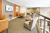 This thoughtfully designed communal space offers a welcoming and sophisticated environment ideal for socializing, small meetings, or quiet relaxation. The interior is anchored by bold burnt-orange accent walls that create striking visual contrast against softer grey tones and textured geometric wallcoverings. Recessed lighting throughout keeps the space bright and well-illuminated, complemented by elegant pendant fixtures that add warmth and dimension.

The layout is open and flowing, featuring multiple distinct seating arrangements that cater to a variety of uses. A lounge area offers comfortable upholstered armchairs paired with a low coffee table, while intimate bistro-style tables with cushioned chairs provide space for casual conversation or individual work. A built-in upholstered bench with a tall decorative back panel and colorful throw pillows adds a boutique-hotel feel to one corner of the space.

The flooring consists of patterned carpet tiles in neutral tones, adding texture underfoot while maintaining a polished, professional aesthetic. Curated artwork, including an expressive abstract canvas with orange, blue, and white tones, enhances the gallery-like atmosphere. A sleek metal and cable railing runs along one edge of the space, suggesting an open interior overlook. Overall, the space balances modern design with comfort and functionality.
