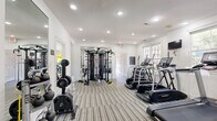 This fitness center offers a thoughtfully designed, single-floor workout space that balances functionality with a clean, modern aesthetic. The interior is finished with crisp white walls, recessed lighting, and a neutral striped flooring that lends a polished, professional feel throughout.

The main workout area is lined with Landice treadmills and elliptical trainers positioned along the windowed wall, allowing natural light to fill the space while offering outdoor views during workouts. Wall-mounted televisions are conveniently placed for entertainment. A robust multi-functional cable and strength training system anchors one end of the room, accommodating a full range of resistance exercises.

A dedicated stretching and group fitness alcove houses spin bikes, exercise mats, resistance bands, medicine balls, stability balls, and foam rollers, all neatly organized on a multi-tier storage rack. Free weights, kettlebells, and a barbell rack round out the strength training offerings. Large wall mirrors are strategically placed to enhance the sense of space and support proper form during exercise.

The facility features multiple access points and is well-ventilated, creating a comfortable environment for varied workout routines. The overall ambiance is bright, open, and inviting, making this fitness center a comprehensive amenity for any level of fitness enthusiast.