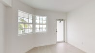 Located at 710 Seventh St in the heart of Miami Beach, FL, this compact yet efficiently designed single-floor space offers a clean, modern aesthetic throughout. The interior is defined by an all-white palette, light-toned large-format tile flooring, and recessed lighting that collectively create a bright and airy atmosphere.

The layout flows from a sunlit main room featuring a curved bay window with multi-pane glass, framing views of swaying palm trees and the surrounding Miami Beach streetscape. Natural light pours generously through multiple windows, including louvered-style upper windows in one of the rooms, casting warm geometric light patterns across the walls and floors.

The kitchen is sleek and contemporary, fitted with high-gloss white cabinetry extending to the ceiling, a flush induction cooktop, stainless steel refrigerator, built-in oven, and a stainless undermount sink — all set against clean white countertops. A wall-mounted ductless air conditioning unit provides climate control in the living area.

A corridor connects the rooms, with a compact bathroom featuring a white vanity and recessed ventilation. The overall design emphasizes clean lines, minimalism, and functionality, making efficient use of the available footprint. This well-lit, move-in-ready space embodies a refined, modern sensibility well-suited to the vibrant Miami Beach locale.