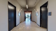 This thoughtfully designed single-floor space masterfully combines historic architectural elements with contemporary finishes to create a refined and functional environment. The entry corridor sets an immediate tone with polished concrete floors, track and sconce lighting, and framed black-and-white photography along neutral gray walls. Dark-framed double glass doors open to the main areas, where the aesthetic shifts dramatically to warm hardwood flooring and elevated ceilings adorned with detailed crown molding.

A signature teal wainscoting runs consistently throughout the main rooms, anchoring the color palette and lending a cohesive, upscale character to the space. The lounge area features expansive industrial-style multi-pane windows that flood the room with natural light and offer sweeping outdoor views, complemented by a leather sofa, curated décor, and a bar-height counter with seating along the window line.

The kitchen is a standout feature, outfitted with sage-toned cabinetry, open shelving, a long communal island with bar stools, and an industrial-style pendant chandelier. A dedicated conference room includes a wood table, leather chairs, a wall-mounted display, and a large window wall with panoramic views. Throughout, the blend of historic detailing and modern appointments creates a polished, welcoming atmosphere.