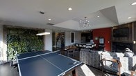 This single-floor space is thoughtfully designed to blend recreation, dining, and relaxation into one cohesive environment. At the forefront is a full-sized ping-pong table, establishing the space's playful, social character. A modern flat pendant light hangs overhead, complementing the recessed lighting throughout the ceiling.

A striking living wall of lush greenery lines one side of the space, adding a natural, vibrant element that contrasts with the sleek, contemporary finishes. Sliding glass barn doors separate an adjacent room, offering flexible use of the layout while maintaining an open feel.

The dining area features a mid-century style table and chairs, positioned beneath a sputnik-style chandelier that adds a retro-modern touch. Adjacent to the dining zone is a well-appointed kitchen with dark cabinetry, stainless steel appliances, and a long bar-height counter with seating — ideal for casual gatherings.

A Starbucks station is integrated near the kitchen, suggesting a commercial or communal-use environment. A bold red accent wall anchors the kitchen area visually. Patterned lounge seating rounds out the space, creating a comfortable retreat within this multifunctional layout.