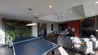 This single-floor space is thoughtfully designed to blend recreation, dining, and relaxation into one cohesive environment. At the forefront is a full-sized ping-pong table, establishing the space's playful, social character. A modern flat pendant light hangs overhead, complementing the recessed lighting throughout the ceiling.

A striking living wall of lush greenery lines one side of the space, adding a natural, vibrant element that contrasts with the sleek, contemporary finishes. Sliding glass barn doors separate an adjacent room, offering flexible use of the layout while maintaining an open feel.

The dining area features a mid-century style table and chairs, positioned beneath a sputnik-style chandelier that adds a retro-modern touch. Adjacent to the dining zone is a well-appointed kitchen with dark cabinetry, stainless steel appliances, and a long bar-height counter with seating — ideal for casual gatherings.

A Starbucks station is integrated near the kitchen, suggesting a commercial or communal-use environment. A bold red accent wall anchors the kitchen area visually. Patterned lounge seating rounds out the space, creating a comfortable retreat within this multifunctional layout.
