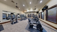 Located at 3290 Dewar Drive in Rock Springs, Wyoming, this fitness center offers a thoughtfully designed exercise space that accommodates a comprehensive range of workout equipment. The single-room facility features vaulted ceilings that lend a sense of openness and volume, complemented by recessed lighting and multiple ceiling fans that ensure a comfortable environment during workouts.

The exercise room is equipped with a variety of cardio machines, including treadmills, ellipticals, and a recumbent bike, all from Spirit fitness equipment. Strength training is well-represented with multiple cable and weight machines positioned on protective rubber floor mats, providing dedicated zones for resistance-based exercise. The neutral-toned carpet tiles throughout the space create a clean, functional aesthetic.

A large wall-mounted mirror spans one side of the room, visually expanding the space and allowing users to monitor their form. Multiple wall-mounted flat-screen televisions are positioned for easy viewing from various workout stations. Dark wood-trimmed windows with slatted blinds line the walls, allowing natural light to filter in while maintaining privacy. The warm, neutral wall tones and dark wood accents throughout give the fitness center a polished, welcoming appearance suited for daily use.