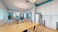 Located at 8485 Hammocks Boulevard in Miami, FL, this thoughtfully designed single-floor communal space delivers a cohesive coastal aesthetic throughout its well-appointed rooms. The interior is defined by a crisp white and soft teal color palette, light wood-plank flooring, and abundant natural light streaming through multiple windows and glass-paned French doors that open to an outdoor area framed by tropical palms.

The open lounge area anchors the space with a comfortable sectional sofa, woven accent chairs, a circular gold pendant light, and large-scale ocean-themed artwork. A decorative lattice wood wall panel adds architectural texture and visual interest. The adjoining dining area features a long natural wood table with cane-back chairs accented in navy, illuminated by a sleek linear gold pendant fixture, and complemented by a wood sideboard and outdoor access via sliding glass doors.

The kitchenette is equipped with white shaker cabinetry, a stainless steel refrigerator, and ample counter space, seamlessly connecting to the dining and lounge zones. Two private office nooks provide focused workspaces, each featuring a built-in wood desk, vertical slatted wood paneling, nature-inspired wallpaper, and a window for natural light — ideal for individual productivity within a shared environment.