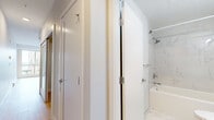 Located on the fourth floor at 2540–2590 Lonsdale Avenue in North Vancouver, this one-bedroom, one-bathroom unit offers a thoughtfully designed layout making excellent use of its compact footprint. Light hardwood flooring runs throughout the space, lending warmth and continuity from room to room.

The open-concept living and kitchen area creates an airy, welcoming atmosphere. The kitchen is neatly integrated into the space, featuring sleek blue-grey lower cabinetry, crisp white upper cabinets, stainless steel appliances including a refrigerator, dishwasher, and built-in microwave, all complemented by a clean white countertop and single-basin sink.

The living area is bathed in natural light, flowing seamlessly toward the bedroom, which is highlighted by large sliding glass doors and floor-to-ceiling windows that frame lush, tree-lined outdoor views — a tranquil connection to the natural surroundings of North Vancouver. A baseboard heating system ensures year-round comfort.

The bathroom is finished to a high standard, featuring marble-effect porcelain tiling on both the floor and walls, a soaker tub with overhead shower, an undermount sink set into a modern vanity with ample counter space, and a large mirror. A hallway connects the main living spaces, completing this well-appointed and efficiently designed home.